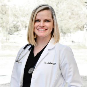 Doctor with blonde hair wearing a lab coat and stethascope smiling while standing in front of a window